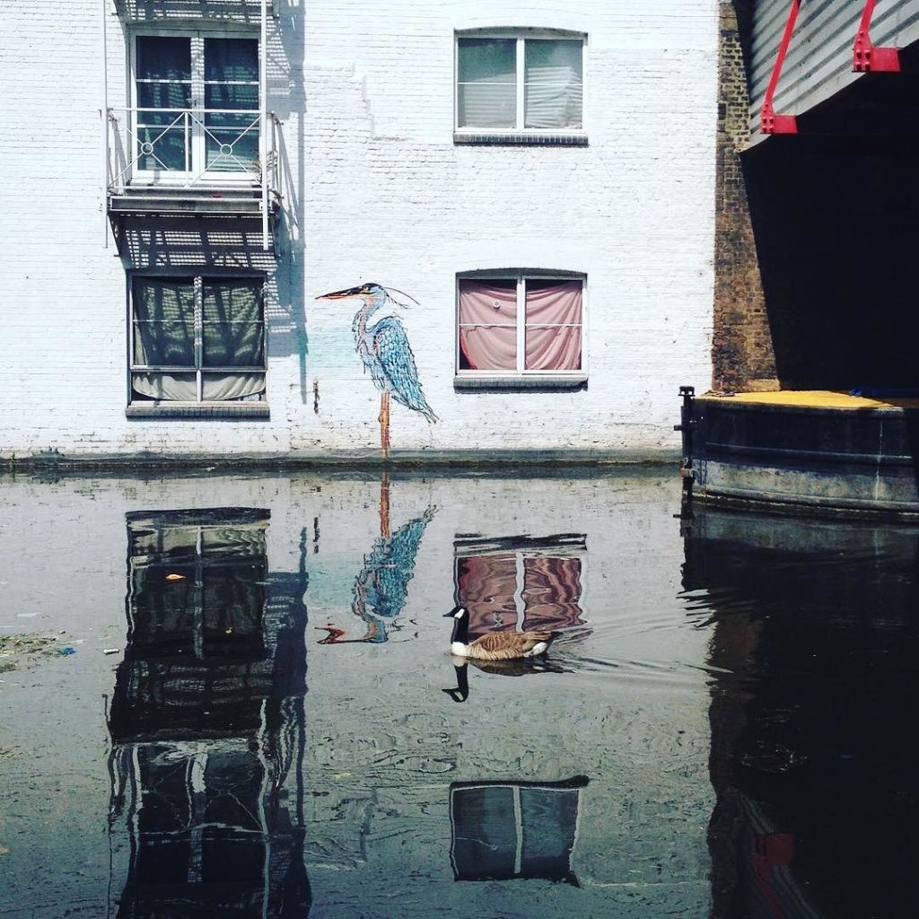 Painting of a heron above the waterline on the side of a building next to the water, with a Canada goose about to cross its reflection.